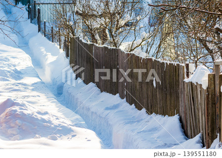 Deep snow-lined country path winds by a rustic fence on a bright winter day. Pristine white drifts create a narrow, inviting trail through the tranquil rural landscape 139551180
