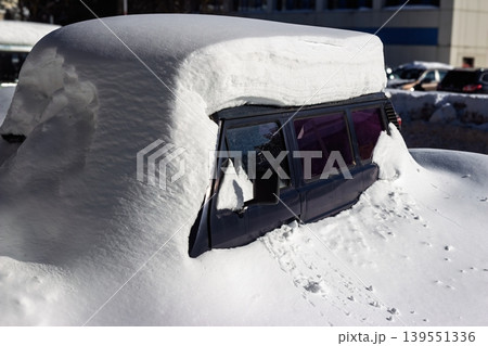 Deep snow blankets a parked vehicle, its windows reflecting the winter sky. The vehicle is almost completely buried, showcasing nature's power and winter's extreme conditions Deep snow blankets a parked vehicle, its windows reflecting the winter sky. The vehicle is almost completely buried, showcasing nature's power and winter's extreme conditions 139551336