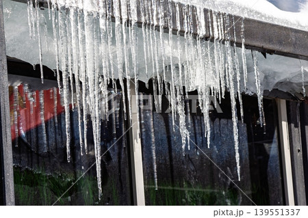 Sparkling melting icicles hang from a roof edge, glistening in sun. Bright, translucent ice formations drip, hinting at the spring thaw. Dynamic winter scene 139551337