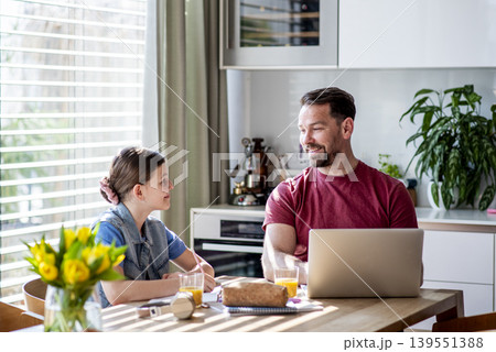 Daughter studying at table as dad works remotely on laptop nearby. Daughter studying at table as dad works remotely on laptop nearby. 139551388