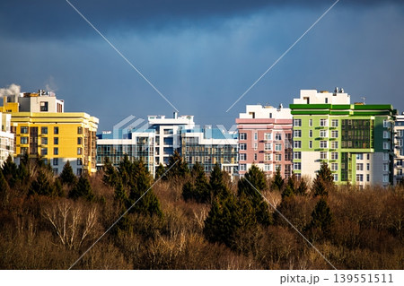 Bright, modern apartment blocks in striking colors peek above a lush forest canopy, showcasing urban development against a dramatic blue sky Bright, modern apartment blocks in striking colors peek above a lush forest canopy, showcasing urban development against a dramatic blue sky 139551511