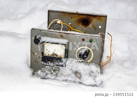 Weathered Soviet-era electronic device, an ammeter and wires exposed, half-buried in fresh winter snow. A defunct relic of industrial tech, forgotten in the cold 139551522