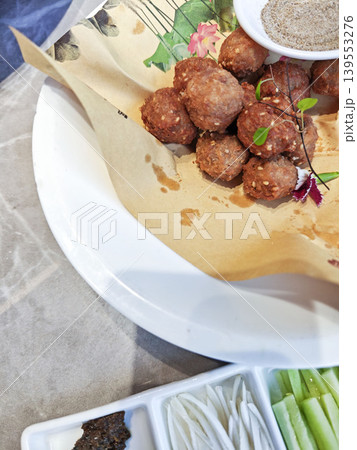 Plate of fried meatballs garnished with herbs and served with a small bowl of seasoning on a decorative paper, accompanied by sliced vegetables on a separate dish 139553276