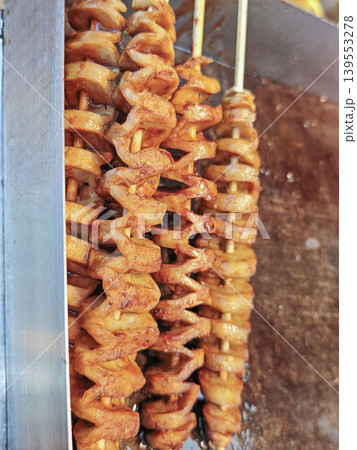 Spiral-cut fried potato skewers seasoned with spices displayed on a metal grill at a street food stall in an outdoor market setting Spiral-cut fried potato skewers seasoned with spices displayed on a metal grill at a street food stall in an outdoor market setting 139553278