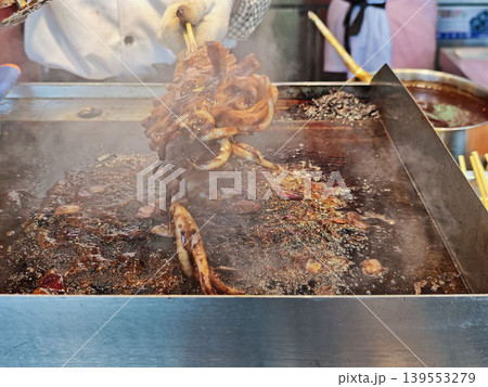 The chef holds a whole squid on a skewer over a sizzling grill, from which steam rises, demonstrating the cooking process amid the lively atmosphere of a street food scene The chef holds a whole squid on a skewer over a sizzling grill, from which steam rises, demonstrating the cooking process amid the lively atmosphere of a street food scene 139553279