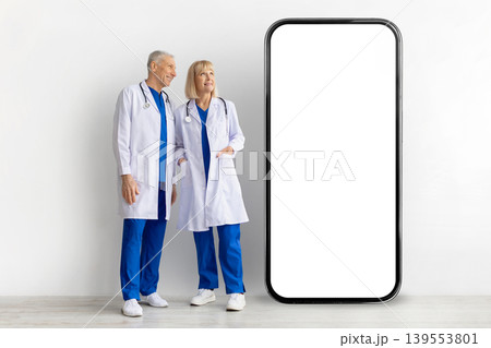 Two mature doctors in lab coats standing beside giant blank smartphone screen 139553801