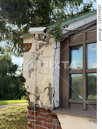Security camera mounted on weathered building corner amidst greenery and cracked walls Security camera mounted on weathered building corner amidst greenery and cracked walls 139554887