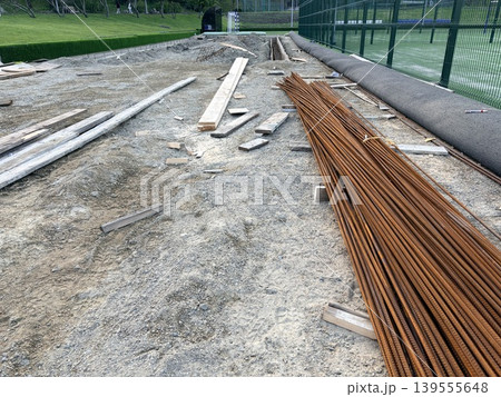an active construction site situated directly beside a green sports field. A strip of prepared earth and gravel is scattered with wooden planks, beams, and construction debris. an active construction site situated directly beside a green sports field. A strip of prepared earth and gravel is scattered with wooden planks, beams, and construction debris. 139555648