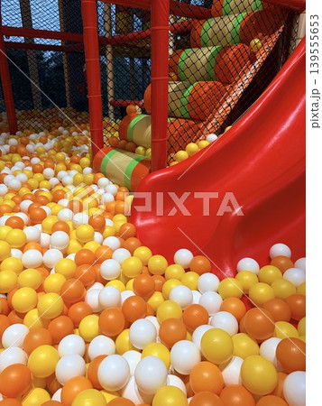 a vibrant section of an indoor soft-play area, focusing on a glossy red plastic slide that curves downward into a densely packed ball pit. 139555653