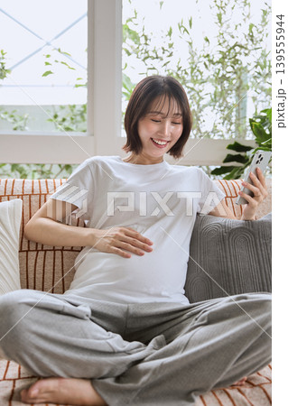 ソファでスマホを見ながら笑顔でお腹を触る妊婦 ソファでスマホを見ながら笑顔でお腹を触る妊婦 139555944