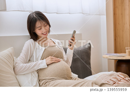 ベッドでスマートフォンを見ながらお腹に手を当てる笑顔の妊婦 139555959