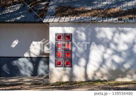 Exterior view of oriental grey and red Chinese style windows on white wall of resident building. The sunlight shines through trees and causes shadows on Chinese style house. Exterior view of oriental grey and red Chinese style windows on white wall of resident building. The sunlight shines through trees and causes shadows on Chinese style house. 139556480