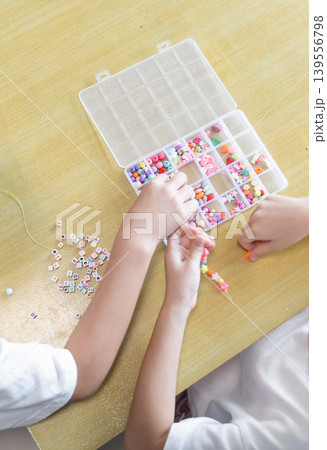 Children thread colorful beads on elastic string at a wooden table. Small hands make a bracelet from a sorted craft box. 139556798