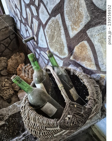 Several bottles of wine in a wicker vintage basket covered in dust and cobwebs. A wall with mosaic elements Several bottles of wine in a wicker vintage basket covered in dust and cobwebs. A wall with mosaic elements 139556932