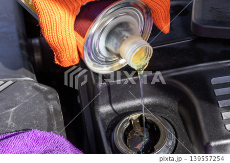A man is pouring oil into the engine of a car while working in a garage during the daytime. The car parts and tools are visible A man is pouring oil into the engine of a car while working in a garage during the daytime. The car parts and tools are visible 139557254