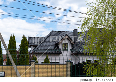 A house stands with a gray roof and tall trees around it. The sky is partly cloudy, and the fence surrounds the yard A house stands with a gray roof and tall trees around it. The sky is partly cloudy, and the fence surrounds the yard 139557284