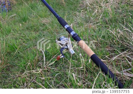 Fishing rod with a golden spinning reel and fishing line in green grass on a riverbank. Concept of outdoor recreation and hobbies. 139557755