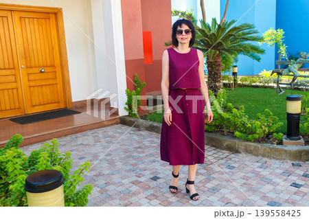 A woman who owns a large house stands near the front door against the backdrop of a green garden 139558425