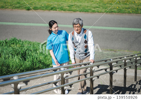 公園の階段を登るシニア男性と付き添いで介助する介護士の女性 公園の階段を登るシニア男性と付き添いで介助する介護士の女性 139558926
