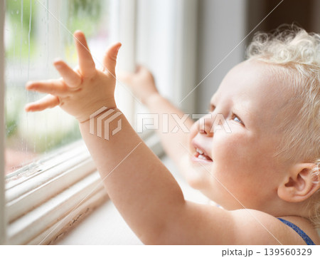 Baby, rain and happy child by window, curious and smile alone with wet weather. Young kid, glass and dream of blonde toddler thinking of childhood, cute girl playing and watch for learning at house 139560329