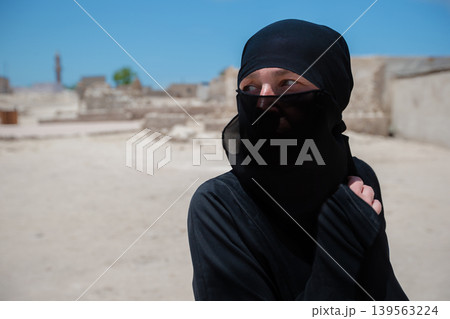 A woman in a black Arabic outfit stands in an ancient archaeological place under a clear blue sky 139563224