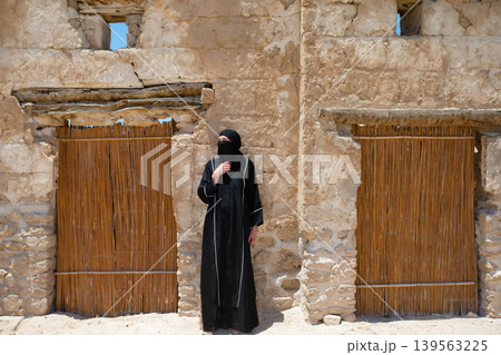 Woman in traditional attire stands beside rustic adobe structure in desert at midday 139563225