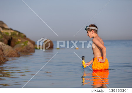 A young boy is immersed in shallow water, focused on his camera as he captures the beauty of the underwater world. The sun sets on the horizon, casting a warm glow. 139563304