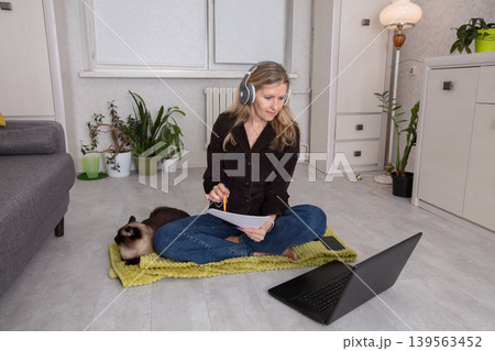 Woman working from home in a cozy space with a cat nearby while sitting on a mat during daylight hours 139563452