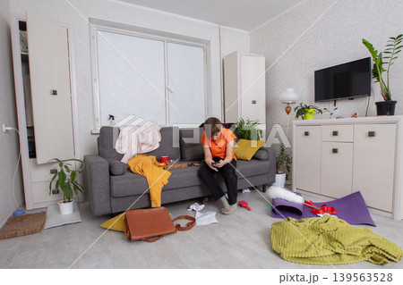 A teenager sits on the couch in a dirty living room, focused on the phone among the mess and colorful decor A teenager sits on the couch in a dirty living room, focused on the phone among the mess and colorful decor 139563528