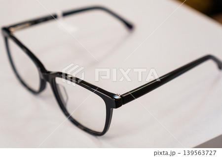 Black plastic eyeglasses frames resting on white surface, minimalist design with clear lenses and glossy finish for visual clarity Black plastic eyeglasses frames resting on white surface, minimalist design with clear lenses and glossy finish for visual clarity 139563727