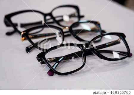 Selective focus black eyeglasses frames arranged on white table surface, optical vision accessory closeup, minimalist composition Selective focus black eyeglasses frames arranged on white table surface, optical vision accessory closeup, minimalist composition 139563760