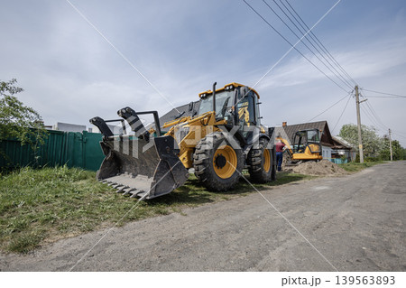 A backhoe loader sits on a dirt road near houses. Workers prepare ground and move dirt. Power lines run along the side of the road under a blue sky 139563893