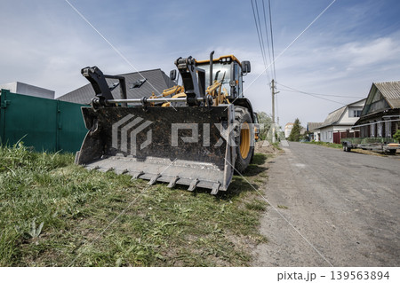 A front loader sits parked on a dirt road beside houses in a small residential area. Sunlight shines on the scene in clear weather A front loader sits parked on a dirt road beside houses in a small residential area. Sunlight shines on the scene in clear weather 139563894