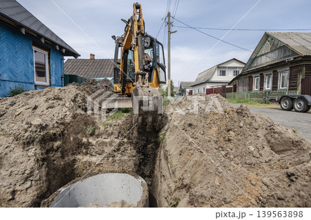 Workers operate a digger to create a trench in a rural setting. Nearby houses and utility poles are visible in the background under an overcast sky Workers operate a digger to create a trench in a rural setting. Nearby houses and utility poles are visible in the background under an overcast sky 139563898