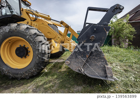 A loader is parked in a yard, ready to dig soil and lift materials. The machine sits near a house with green grass around it on a cloudy day 139563899