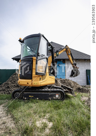 A small excavator is digging soil at a construction site. The machine is next to a building that is being developed. There are dirt piles around the area 139563903