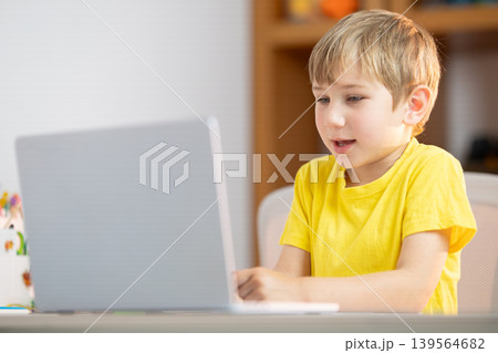 Young boy works on laptop at a table during daytime in a simple room while focusing on his task Young boy works on laptop at a table during daytime in a simple room while focusing on his task 139564682