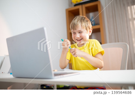 Young boy smiles while using laptop at home during afternoon learning session with colorful pens and paper Young boy smiles while using laptop at home during afternoon learning session with colorful pens and paper 139564686