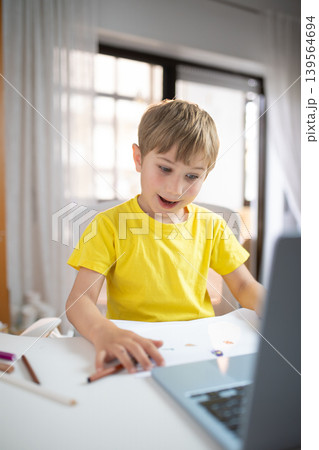 Young child with blonde hair sits at table using laptop while engaged in creative activity at home during daytime 139564694