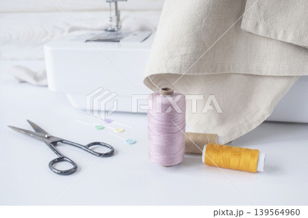 Sewing supplies on a white table with a sewing machine in the background. Sewing machine with fabric and sewing accessories on a white table. 139564960