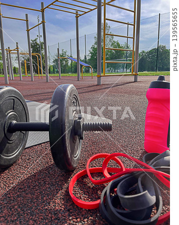 Outdoor fitness equipment on sports ground, barbell resistance bands and water bottle at calisthenics park, strength training and healthy lifestyle. Outdoor fitness equipment on sports ground, barbell resistance bands and water bottle at calisthenics park, strength training and healthy lifestyle. 139565655