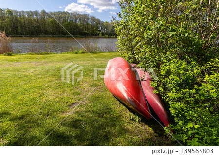Red canoe resting on green lakeside grass near forest and water. Red canoe resting on green lakeside grass near forest and water. 139565803
