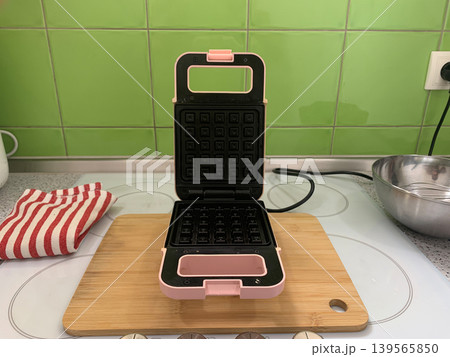 High angle centered view of an open pink waffle maker on a wooden board with a striped towel. High angle centered view of an open pink waffle maker on a wooden board with a striped towel. 139565850