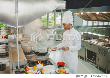 Culinary student cooks with wok and steam in modern professional kitchen. Concept of food preparation, education, skill training, and gastronomy learning Culinary student cooks with wok and steam in modern professional kitchen. Concept of food preparation, education, skill training, and gastronomy learning 139567564