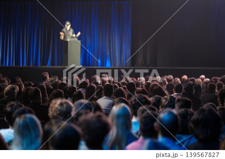 Audience watching speaker on stage at conference presentation with blue curtain and auditorium crowd Audience watching speaker on stage at conference presentation with blue curtain and auditorium crowd 139567827