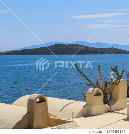 Looking over a traditional building roof with chimneys and growing cactus, featuring the calm blue sea and distant hills 139569132