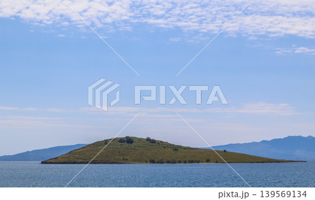 Small green island floating on tranquil blue sea, distant mountains on the horizon, bright sky with light clouds Small green island floating on tranquil blue sea, distant mountains on the horizon, bright sky with light clouds 139569134