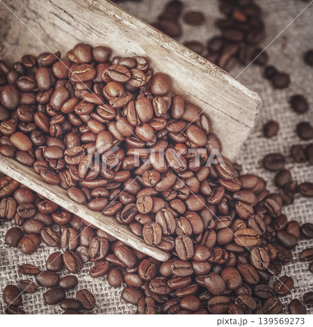 Roasted coffee beans fill wooden scoop on burlap surface, coffee beans scattered around for beverage packaging. Natural background of coffee beans, design poster 139569273