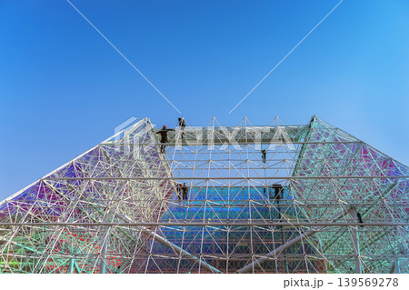 Workers assemble metal structure on geometric glass facade under blue sky with wide. Concept of structure, architecture, construction, engineering, teamwork, urban development. Copy space Workers assemble metal structure on geometric glass facade under blue sky with wide. Concept of structure, architecture, construction, engineering, teamwork, urban development. Copy space 139569278