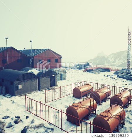 Nestled in a snowy terrain, a collection of red and gray buildings stands near large fuel tanks. The scene is serene, with rocky mountains in the backdrop under a pale sky. Nestled in a snowy terrain, a collection of red and gray buildings stands near large fuel tanks. The scene is serene, with rocky mountains in the backdrop under a pale sky. 139569820